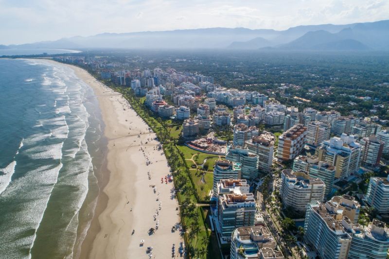 Vista aérea de uma longa praia com diversos edifícios residenciais à beira-mar e montanhas cobertas por vegetação ao fundo.