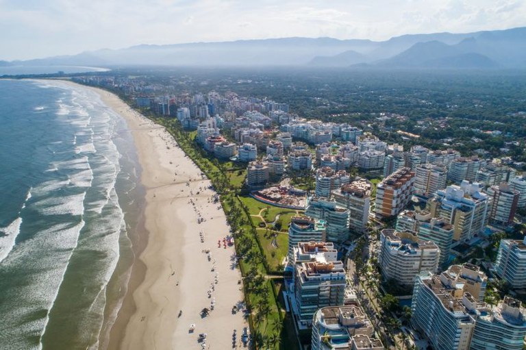 Vista aérea de uma longa praia com diversos edifícios residenciais à beira-mar e montanhas cobertas por vegetação ao fundo.