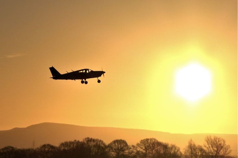 Imagem mostra silhueta de avião de pequeno porte sobrevoando em um céu de tom amarelo, com o sol brilhando ao fundo