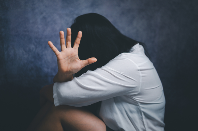 Uma mulher sentada com o rosto escondido e a mão aberta estendida com o sentido de "Pare"