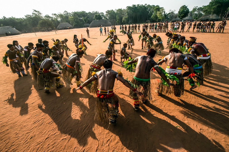 Foto mostra grupo de homens indígenas com trajes tradicionais e cocares coloridos em círculo durante dança em aldeia. Ao fundo, vê-se outros indígenas lado a lado e algumas ocas.