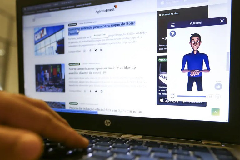 Uma imagem em plano médio e ângulo diagonal mostra a mão em desfoque de uma pessoa digitando no teclado de um notebook da marca HP. A tela do computador exibe o portal de notícias "Agência Brasil", com destaque para uma janela pop-up aberta no canto direito com a ferramenta de acessibilidade "VLibras". Dentro dessa janela, há o avatar digital de um homem jovem de animação 3D, com pele morena, cabelos pretos e camisa azul, posicionado com as mãos unidas à frente do corpo, pronto para traduzir o conteúdo do site para a Língua Brasileira de Sinais.