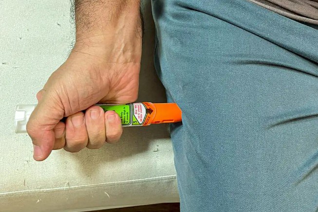Fotografia da mão de um homem aplicando uma caneta de adrenalina em sua própria perna, vestido com uma calça azul.