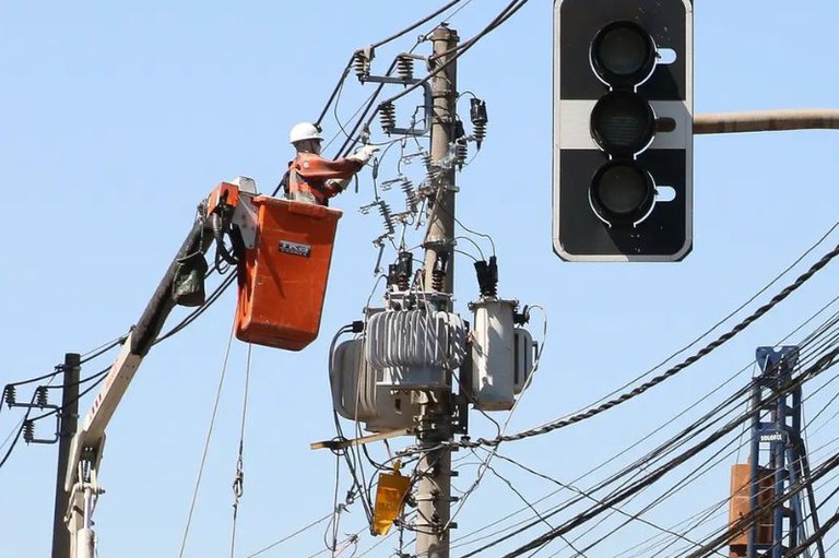 Imagem mostra profissional de empresa concessionária sobre cesto de grua mexendo na fiação em um poste. Em primeiro plano, há um semáforo fora de funcionamento
