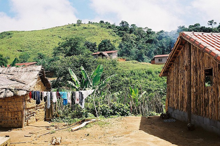 A imagem mostra uma paisagem rural com casas feitas de madeira, barro e telhas. Em primeiro plano, há roupas estendidas em um varal. Ao fundo, vê-se uma área verde com vegetação densa e morros.