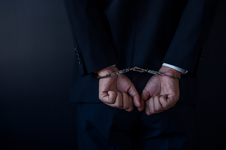 Uma pessoa de costas, vestindo um terno escuro, com as mãos algemadas para trás. A imagem tem um tom sombrio e fundo escuro.