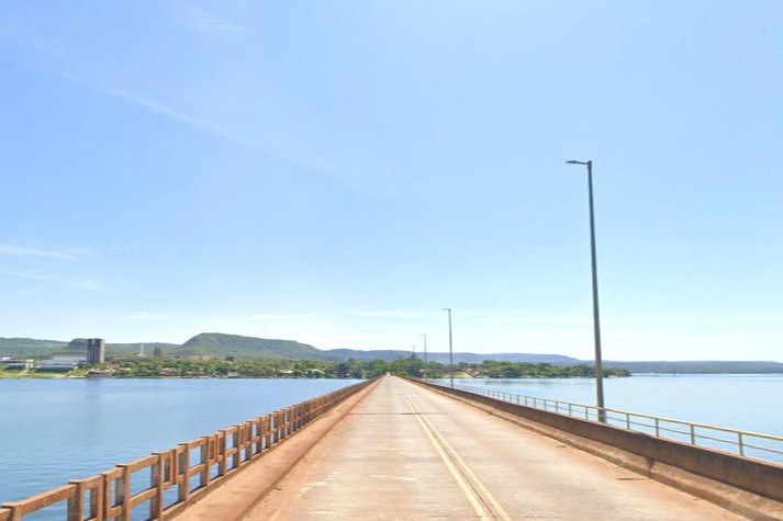 A imagem apresenta uma perspectiva central e profunda de uma ponte rodoviária que cruza o Rio Grande, conectando Rifaina (SP) a Sacramento (MG) sob um céu azul límpido e ensolarado. A estrada, marcada por linhas amarelas desgastadas, conduz o olhar em direção ao horizonte, onde se vê a margem oposta composta por vegetação densa, colinas suaves e algumas edificações isoladas. A estrutura da ponte é ladeada por guardas-corpos distintos: à esquerda, uma proteção de concreto com aberturas retangulares, e à direita, um guarda-corpo metálico acompanhado por altos postes de iluminação que reforçam a escala da travessia sobre as águas calmas e azuladas do reservatório.