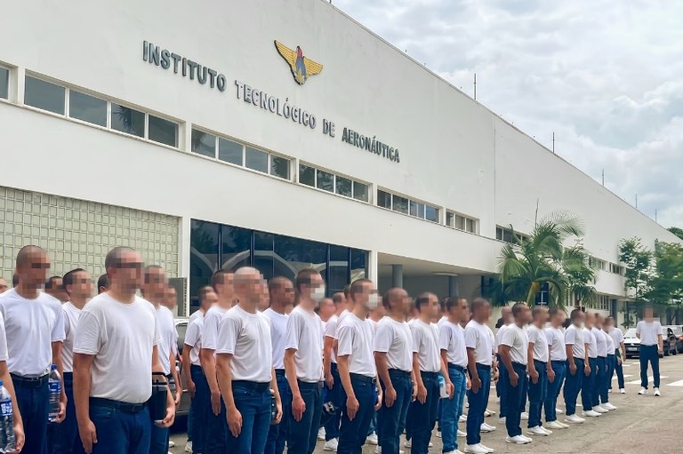 Fotografia colorida, tirada em plano médio e em ângulo ligeiramente lateral, mostrando um grande grupo de jovens recém-ingressos em formação militar ao ar livre. Eles estão posicionados em frente à fachada principal do Instituto Tecnológico de Aeronáutica (ITA).