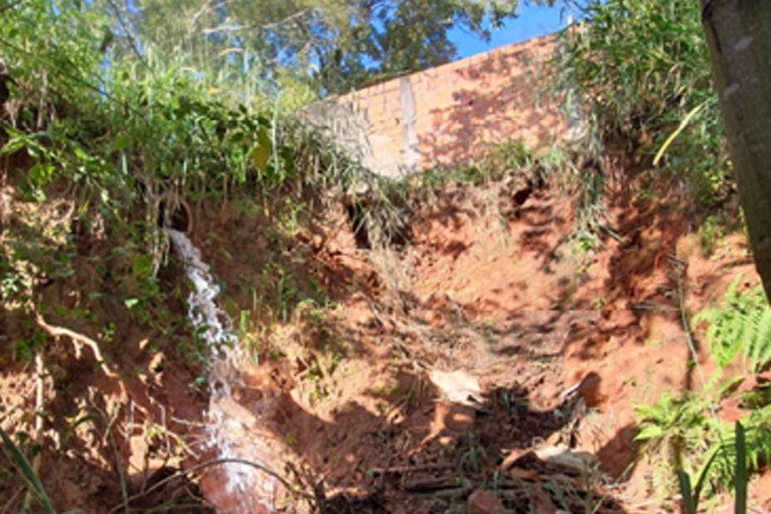 Fotografia de uma tubulação pluvial rompida, ao lado de um muro, despejando água sobre um barranco,que está sofrendo um processo erosivo, cercado por vegetação.