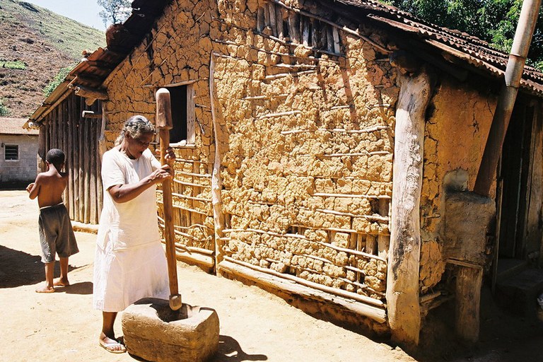 A imagem mostra uma mulher quilombola usando um pilão de madeira em frente a uma casa de taipa. Ela veste roupa branca e está descalça. Ao fundo, um menino sem camisa caminha no terreno.