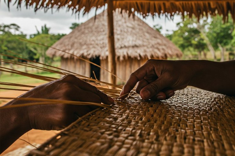 Foto mostra mãos pretas trançando palha. Ao fundo casas com telhado de palha