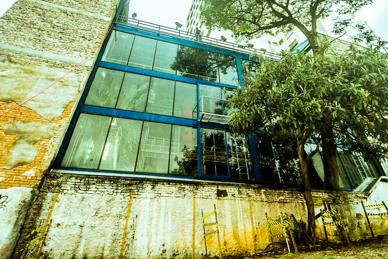 A imagem mostra a fachada de um prédio moderno, possivelmente um teatro, com grandes janelas de vidro azul. Ele está ao lado de uma estrutura mais antiga, de tijolos e reboco descascado, que contrasta com a arquitetura contemporânea. À direita, uma árvore de grande porte cresce perto da construção, adicionando um elemento natural à cena urbana. Fitas de sinalização e detritos no chão sugerem que a área pode estar em reforma ou em desuso. A iluminação e o ângulo da foto dão um ar dramático à cena.