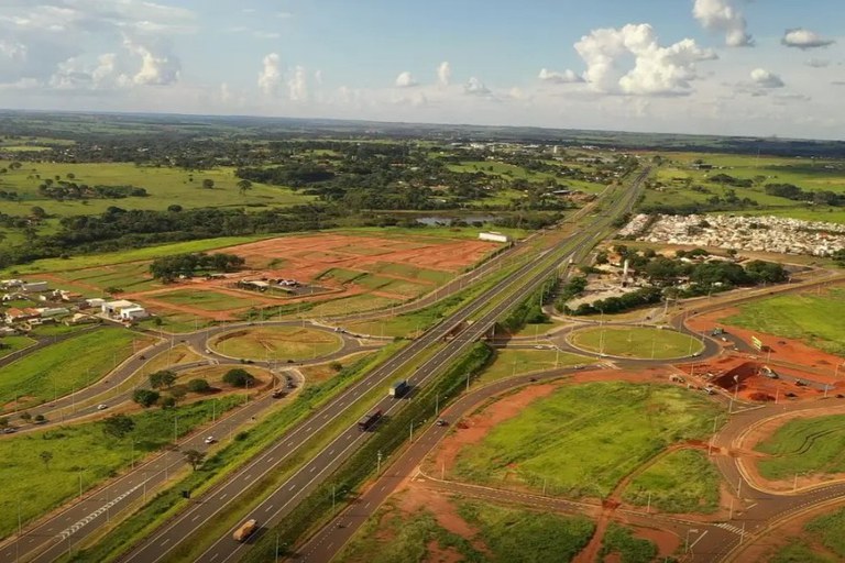 A imagem anexa apresenta uma vista aérea de um trecho da Rodovia BR-153 em São José do Rio Preto, São Paulo, destacando as obras de duplicação e infraestrutura viária em andamento, conforme sugerido pelo nome do arquivo. No centro, a rodovia duplicada se estende pelo cenário, com tráfego de veículos, incluindo caminhões. A via é ladeada por grandes áreas de terraplanagem e desenvolvimento imobiliário (lotes de terra avermelhada), indicando crescimento urbano na região. O design da estrada é complementado por interseções e rotundas (rotatórias) bem definidas, que buscam organizar o fluxo de veículos de e para a rodovia. O ambiente é predominantemente verde, com extensas áreas de pastagem ou vegetação nativa ao redor da área de construção. O céu é parcialmente azul, pontilhado por nuvens brancas e fofas, sugerindo um dia claro e ensolarado.