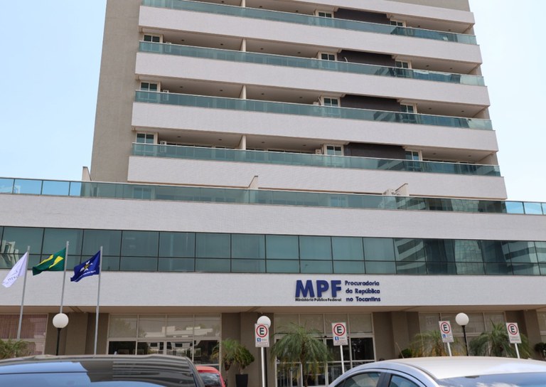 Fotografia em ângulo frontal e de baixo para cima mostrando a fachada do prédio do Ministério Público Federal no Tocantins (MPF). O edifício tem arquitetura moderna com varandas de vidro, fachada bege com cinza e três mastros com bandeiras à esquerda. Na entrada do prédio, destaca-se o logotipo do MPF em azul.
