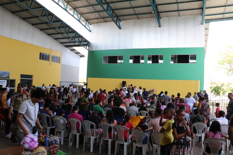 A imagem mostra o pátio de triagem do 2º Pop Rua Jud. Há muitas pessoas reunidas, incluindo adultos e crianças, algumas sentadas em cadeiras plásticas brancas e outras em pé, circulando pelo local. No fundo, há uma parede com pintura em verde e amarelo. Também há exposições de trabalhos manuais e artesanato em uma mesa no canto inferior esquerdo da imagem. O ambiente é coberto, mas bem iluminado, e a estrutura do telhado é metálica com vigas azuis.