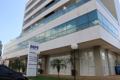 A imagem mostra a fachada da sede do MPF em Palmas, um prédio moderno, com paredes de vidro e estrutura em tons claros. Na frente do prédio há uma placa vertical com a sigla "MPF" (Ministério Público Federal) e o texto "Procuradoria da República no Tocantins". O edifício tem várias janelas espelhadas, algumas plantas ornamentais e postes de iluminação na calçada.