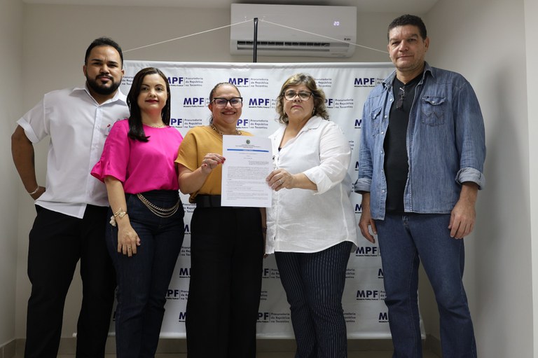 Cinco pessoas (três mulheres e dois homens) posam para uma foto oficial em um ambiente interno. Ao centro, duas mulheres seguram juntas um documento. O grupo está posicionado à frente de um painel branco com o logotipo repetido do Ministério Público Federal, que identifica a unidade como a Procuradoria da República no Tocantins. Acima do painel, há um ar-condicionado split ligado. As pessoas vestem roupas casuais e profissionais, e o clima sugere a formalização de um acordo ou entrega de documento.