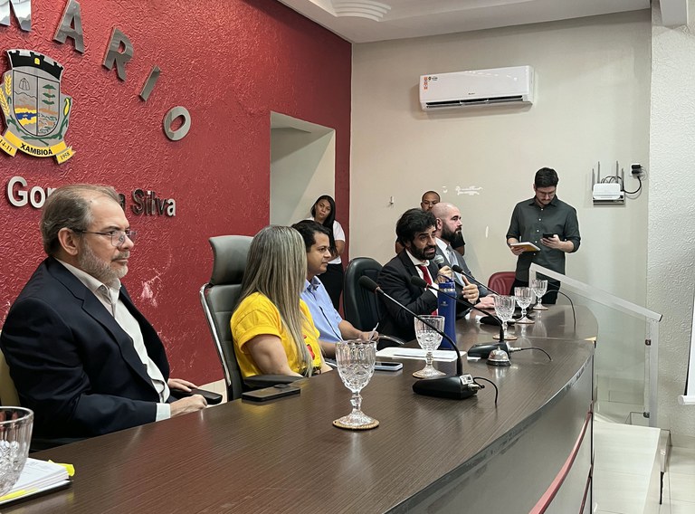 Seis pessoas estão sentadas lado a lado em uma mesa curva de madeira escura, voltada para o público. A parede ao fundo é vermelha com textura e tem o brasão do município de Xambioá (TO), acompanhado do texto “Plenário Governador José da Silva”.