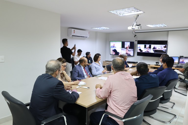 Várias pessoas reunidas em diferentes locais. É possível ver participantes sentados em mesas e outros em frente às câmeras, em diferentes janelas da chamada. Em uma das telas, há uma pessoa falando ou participando de uma reunião, enquanto outras aparecem acompanhando a discussão.