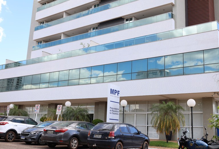 Fotografia da fachada da Procuradoria da República no Tocantins (MPF), mostrando um edifício moderno de vários andares, com fachada branca e amplas janelas de vidro azul refletindo o céu. Na entrada, há uma placa com a identificação do MPF, além de carros estacionados e algumas plantas ornamentais em frente ao prédio.