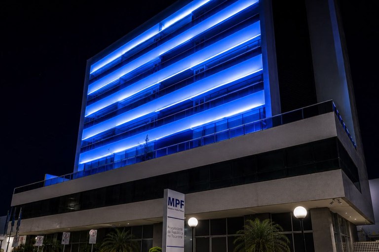 Fotografia noturna da fachada da Procuradoria da República no Tocantins (MPF-TO), com luzes azuis intensas iluminando as varandas dos andares superiores.