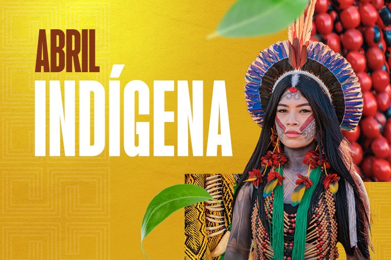 Foto de uma pessoa indígena de costas usando um grande cocar de penas azul-escuro em formato de leque, com três penas amarelas no topo. O cabelo está dividido em duas tranças longas sobre uma roupa preta. Ao fundo, vê-se o teto de palha de uma construção e a folhagem verde de árvores sob a luz do dia.