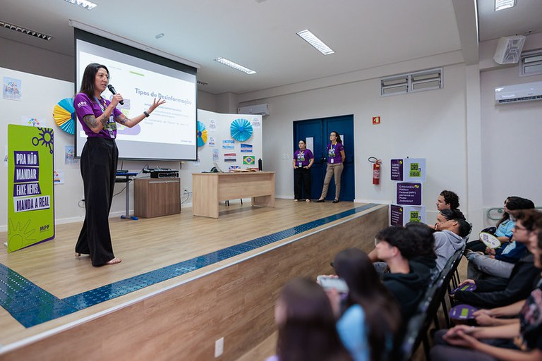 Foto mostra um auditório com alunos sentados nas cadeiras enquanto assistem à palestra de uma mulher em cima de um tablado com um cartaz com texto que diz para não mandar fake news manda a real e ao lado, atrás da palestrante um telão com informações da palestra onde se lê tipos de desinformação; do outro lado do tablado, duas mulheres em pé acompanham a palestra