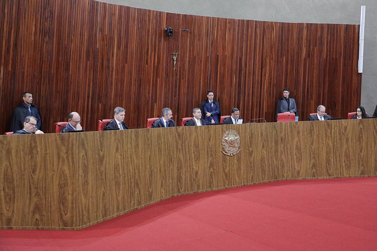 Sessão do Tribunal Superior Eleitoral (TSE) com juízes e ministros vestindo togas e sentados na bancada de madeira. À frente, um púlpito, piso vermelho e a plateia disposta em auditório.
