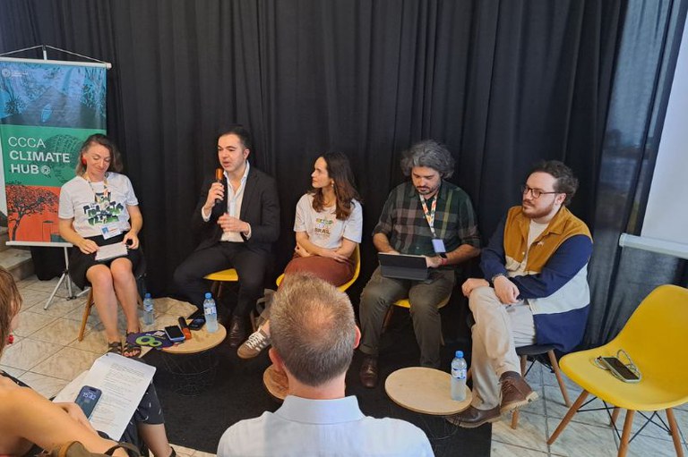 Cinco pessoas participam de um painel de debate em um ambiente interno, sentadas lado a lado em cadeiras amarelas diante de uma cortina preta. Uma delas fala ao microfone enquanto as outras escutam atentamente. À esquerda, há um banner com os dizeres “CCCA Climate Hub”. Na frente, o público acompanha a conversa.