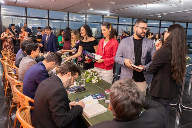 Pessoas se reúnem em uma mesa comprida. Em primeiro plano, um homem autografa um livro, enquanto outros interagem ao redor, com alguns segurando exemplares.