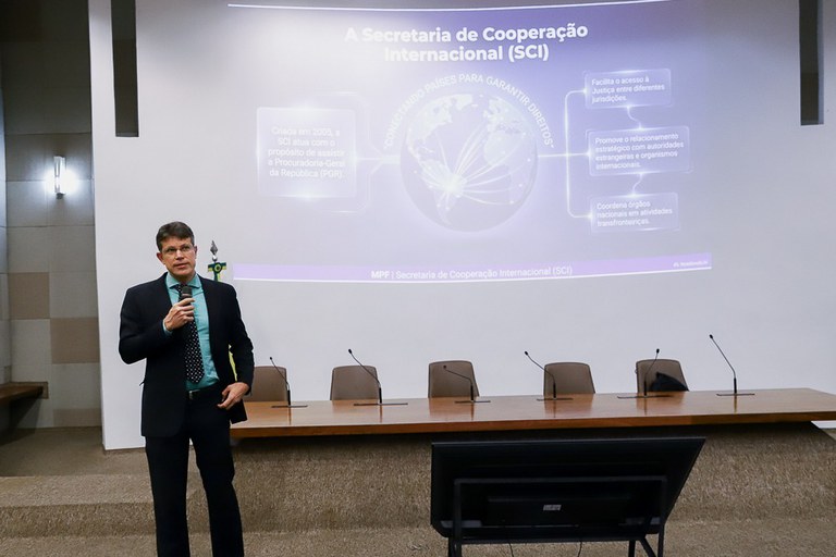 Foto de palestrante num palco, à frente de um telão no qual está projetada uma apresentação com o título Secretaria de Cooperação Internacional