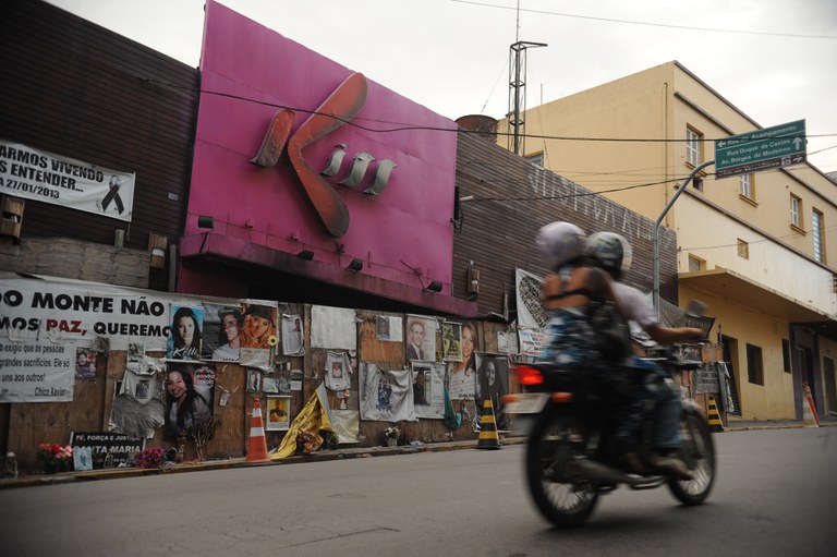 Foto da Boate Kiss, localizada no município de Santa Maria (RS). Há tapumes com fotos de pessoas que morreram no local. Uma moto passa na frente.