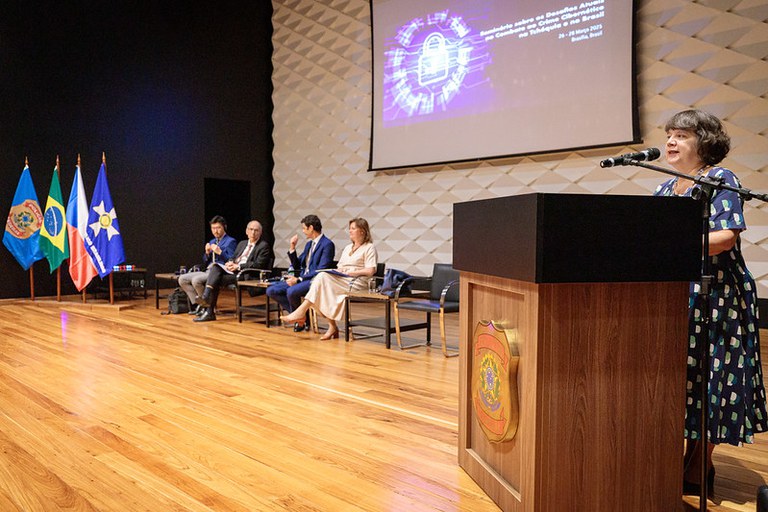 Imagem da representante do MPF falando de pé e os demais palestrantes da abertura sentados ao fundo