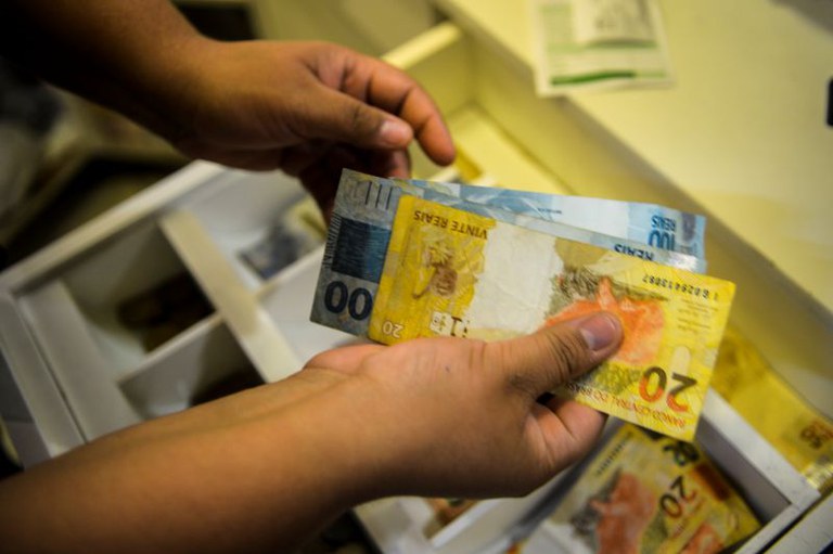 Foto de cédulas de dinheiro. Em primeiro plano, duas mãos seguram duas notas de R$ 100 (cem reais) e uma nota de R$20 (vinte reais). Ao fundo, uma gaveta utilizada por bancários possui outras cédulas.