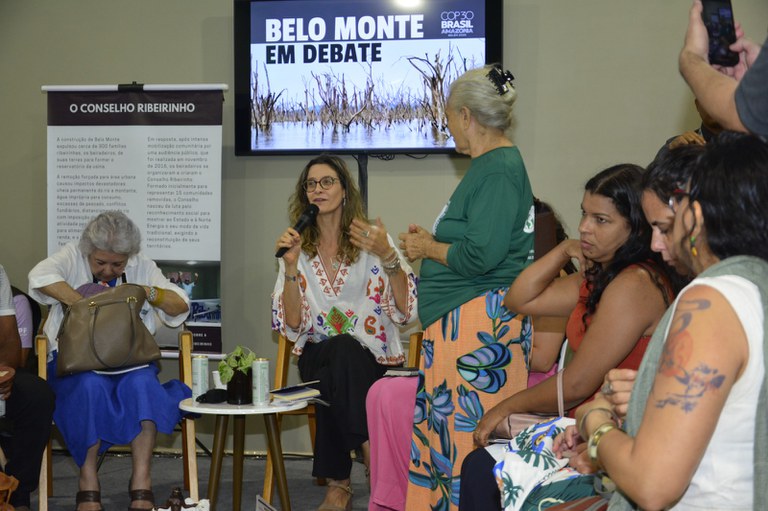 Uma mulher sentada no centro, falando ao microfone durante um debate sobre Belo Monte, exibido em um monitor ao fundo. Várias mulheres assistem, com uma senhora em pé à direita.