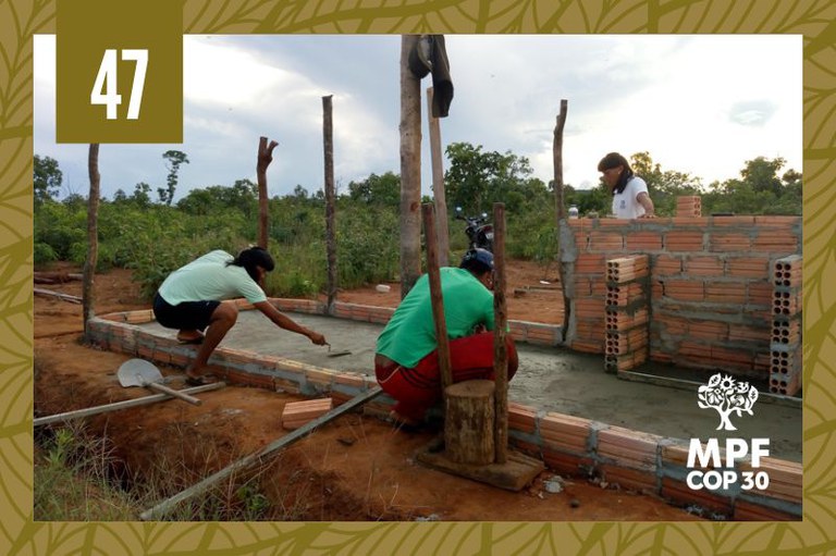 A imagem mostra uma cena de construção em área rural, com três pessoas trabalhando em uma estrutura de tijolos e concreto. Há vegetação ao fundo e estacas de madeira ao redor do local.
