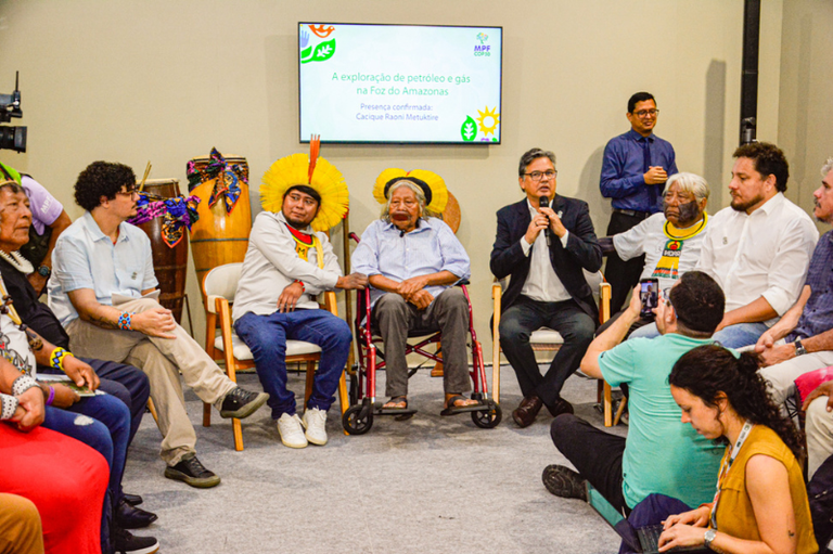 Debate sobre a exploração de petróleo e gás na Foz do Amazonas, com a participação de dois líderes indígenas de cocar e um debatedor falando ao microfone.