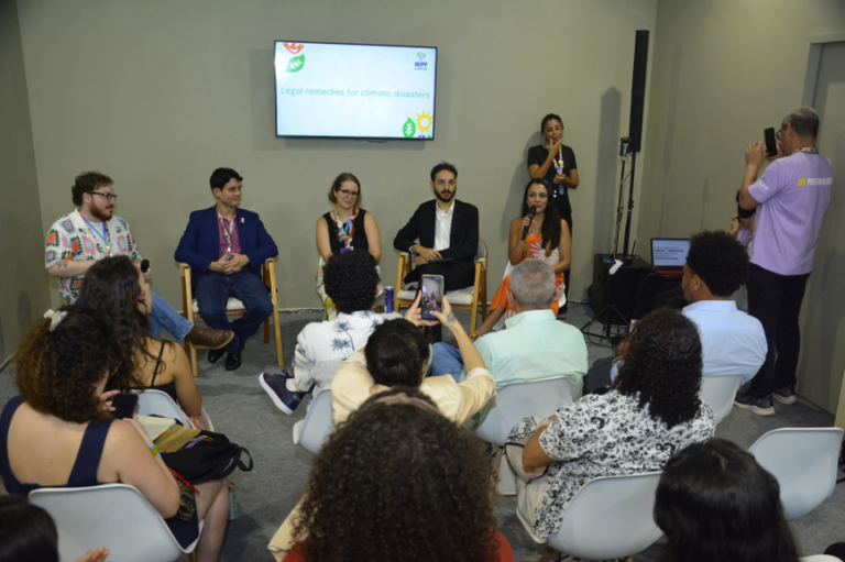 Foto mostra painel de debate sobre "Legal remedies for climate disasters" com cinco participantes sentados em cadeiras e uma debatedora falando ao microfone para uma plateia.