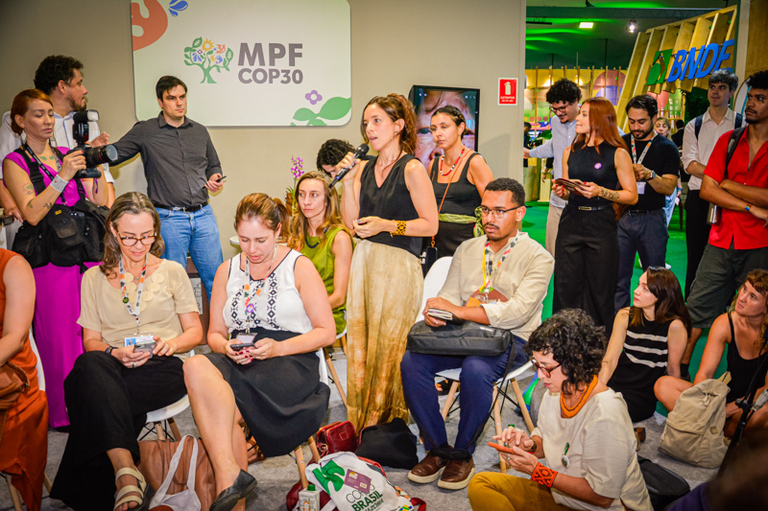 Foto de uma mulher em pé falando em um microfone para uma plateia sentada e em pé em uma reunião no evento MPF COP30.