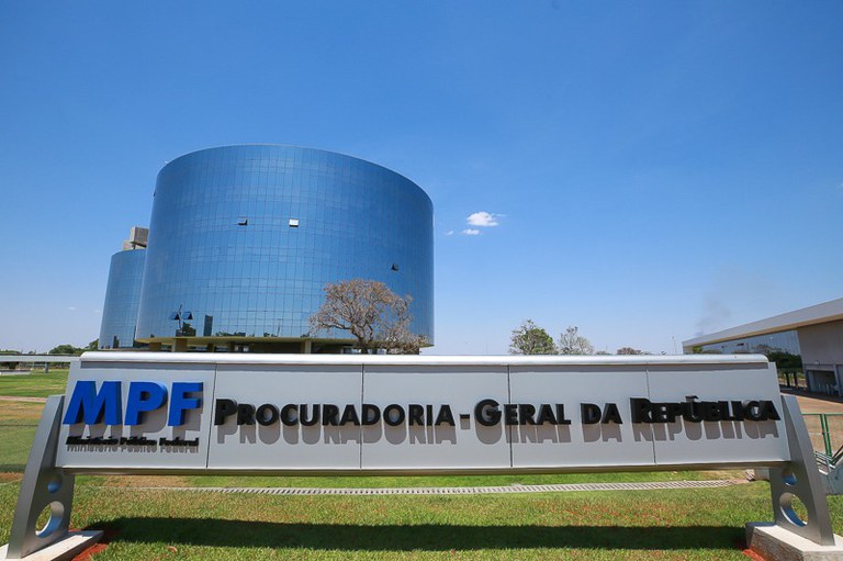 Foto dos prédio da PGR. ME primeiro plano, há uma placa de visualização grande marca com a inscrição da marca do MPF (Ministério Público Federal) em azul e o texto Procuradoria-Geral da República, em preto