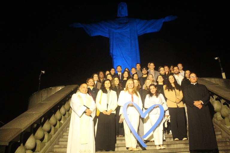 Fotografia noturna mostrando um grupo de cerca de vinte e cinco pessoas posando nas escadarias aos pés do monumento do Cristo Redentor, no Rio de Janeiro. Ao fundo, a estátua do Cristo está iluminada com uma luz azul intensa contra o céu escuro da noite.