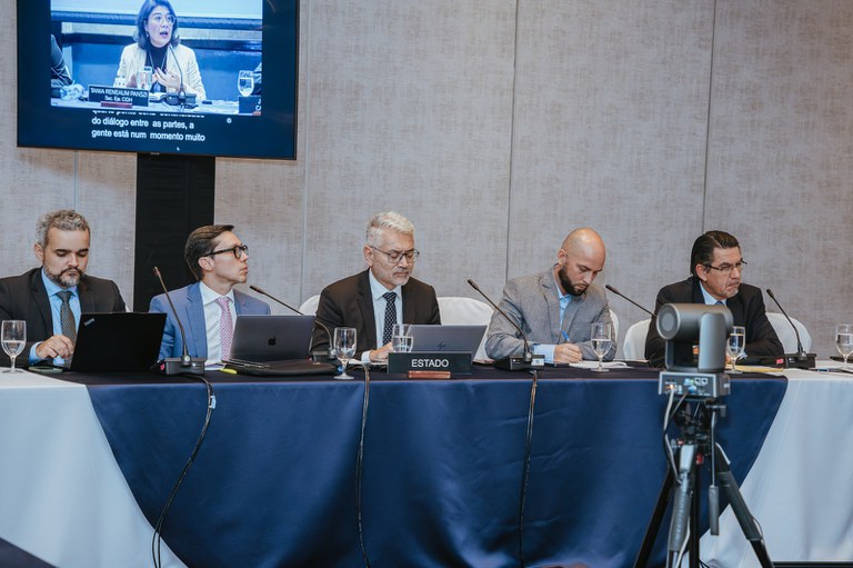 Cinco homens sentados atrás de uma mesa comprida participam de um painel ou reunião formal, cada um com microfone, computadores e copos d’água à frente. Todos usam terno e parecem concentrados, alguns olhando para laptops enquanto outro faz anotações em um bloco. Ao fundo, há um telão exibindo uma mulher falando em outra mesa de debate, e na frente da mesa aparece um equipamento de filmagem voltado para os participantes.