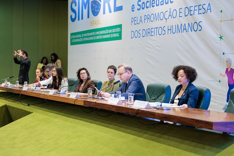 Uma mesa composta por sete pessoas sentadas em uma longa bancada de madeira durante um evento oficial. Ao fundo, um grande banner branco exibe o logotipo "SIMORE BRASIL" .