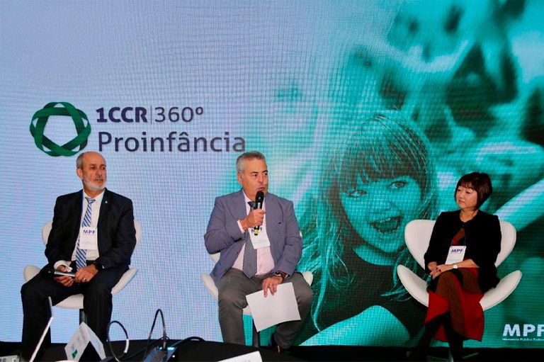 A imagem mostra três pessoas sentadas em cadeiras brancas em um palco, com um grande telão ao fundo.