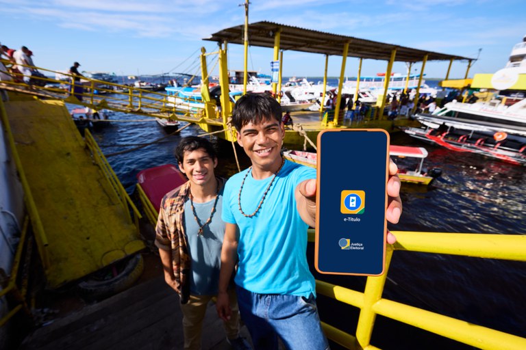 Imagem de dois indígenas segurando e-título em um ambiente próximo a um rio com embarcações ao fundo