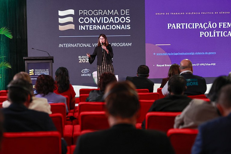 Fotografia de evento em auditório do TSE. Em primeiro plano, vistas de costas, estão várias pessoas sentadas em poltronas vermelhas, acompanhando a apresentação. No centro do palco, em pé, uma mulher de cabelos castanhos na altura dos ombros e óculos fala ao microfone segurando um passador de slides. Ela veste uma blazer preto sobre um vestido longo estampado.

Ao fundo, há painéis em tons de cinza e roxo. No painel da esquerda, lê-se em letras maiúsculas brancas: "PROGRAMA DE CONVIDADOS INTERNACIONAIS" e "INTERNATIONAL VISITORS PROGRAM", acompanhados do logotipo do evento. No painel roxo, à direita, destaca-se o texto: "GRUPO DE TRABALHO VIOLÊNCIA POLÍTICA DE GÊNERO" e o título "PARTICIPAÇÃO FEMININA NA POLÍTICA", além do link institucional do MPF e o nome da palestrante, Raquel Branquinho.