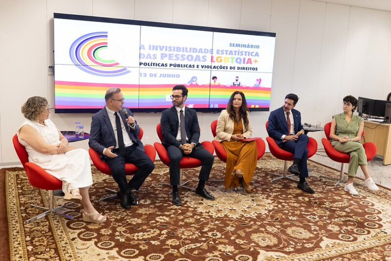A imagem mostra um painel de discussão com cinco pessoas sentadas em poltronas vermelhas sobre um tapete estampado, enquanto um homem fala ao microfone.
Ao fundo, há uma grande tela digital com o título "Seminário: A Invisibilidade Estatística das Pessoas LGBTQIA+ - Políticas Públicas e Violações de Direitos"