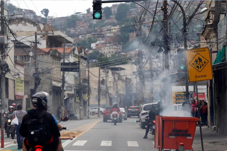 Foto de uma comunidade no Rio de Janeiro durante operação policial, com motos e carros em meio à fumaça