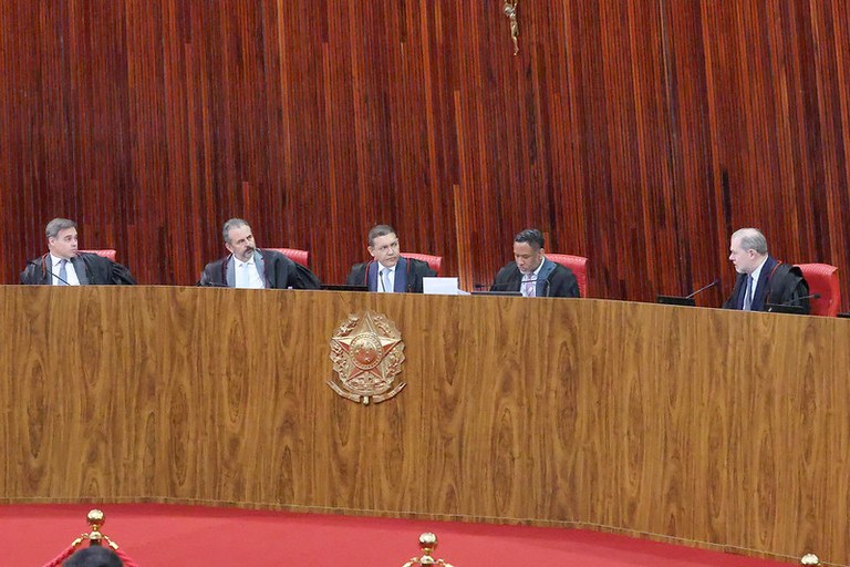 Cinco magistrados de toga sentados atrás de uma bancada curva de madeira com o Brasão da República do Brasil, diante de um painel de madeira com um crucifixo ao fundo.