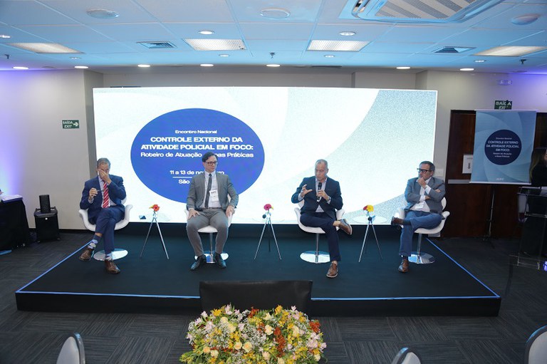 Foto de um painel de debate no Encontro Nacional de Controle Externo da Atividade Policial, em São Paulo. Quatro homens de terno e camisa social estão sentados em cadeiras brancas sobre um palco preto elevado. Eles gesticulam enquanto conversam. Ao fundo, um telão luminoso exibe o título do evento: "Encontro Nacional 'Controle Externo da Atividade Policial em Foco: Roteiros de Atuação e Boas Práticas'". O ambiente é moderno, com iluminação azul e um arranjo de flores na frente do palco.